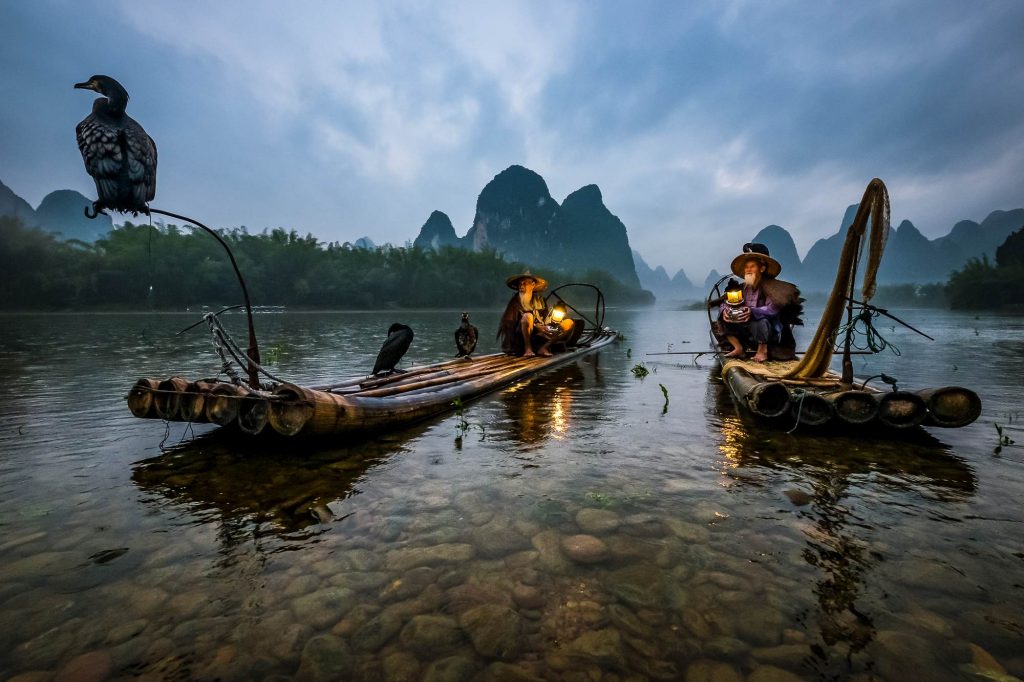 Cormorant fishing is a traditional fishing method in which fishermen use trained cormorants to fish in rivers. Two fishermen are seen as they take rest near the shore.