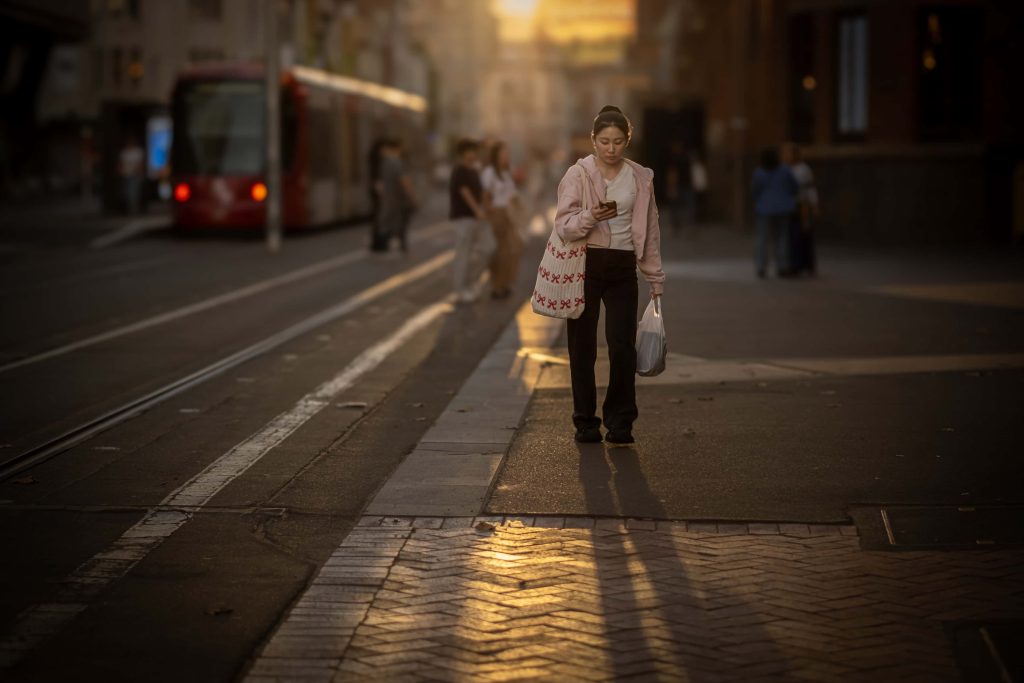 A woman in the foreground, elegantly backlit by golden sunlight, her silhouette framed against the tram tracks and city backdrop. A serene moment of solitude within the urban landscape.