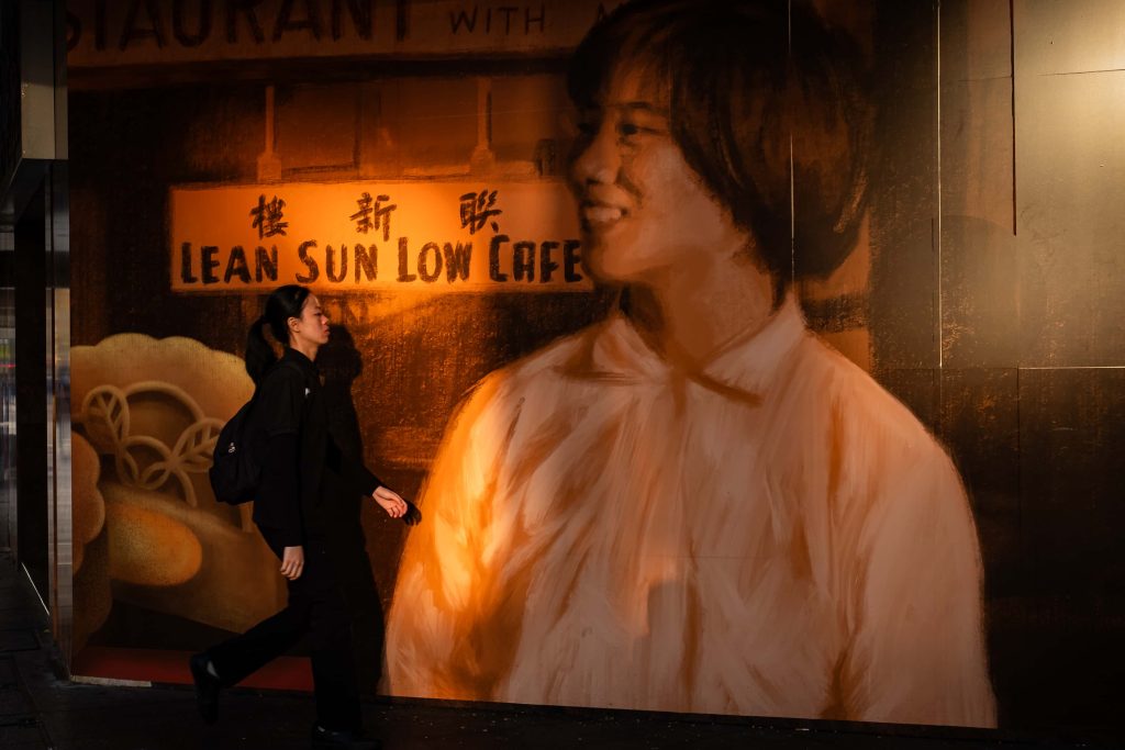 A solitary figure in black steps swiftly past a glowing cafe mural, contrasting fleeting motion with the timeless warmth of street art.